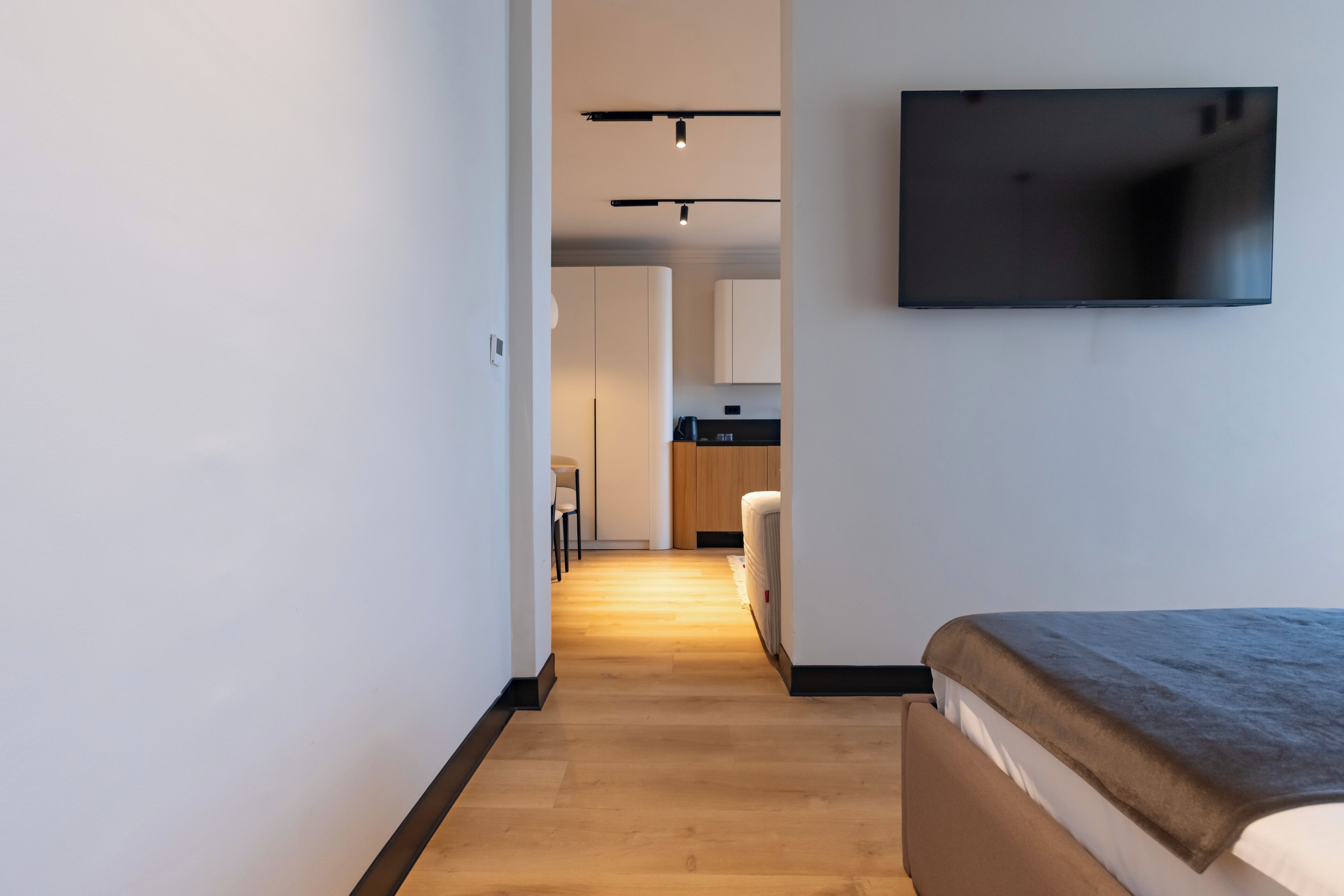 Een moderne hotelkamer met een houten vloer, een bed, een flatscreen televisie aan de muur en een keukenruimte zichtbaar door een open deur.