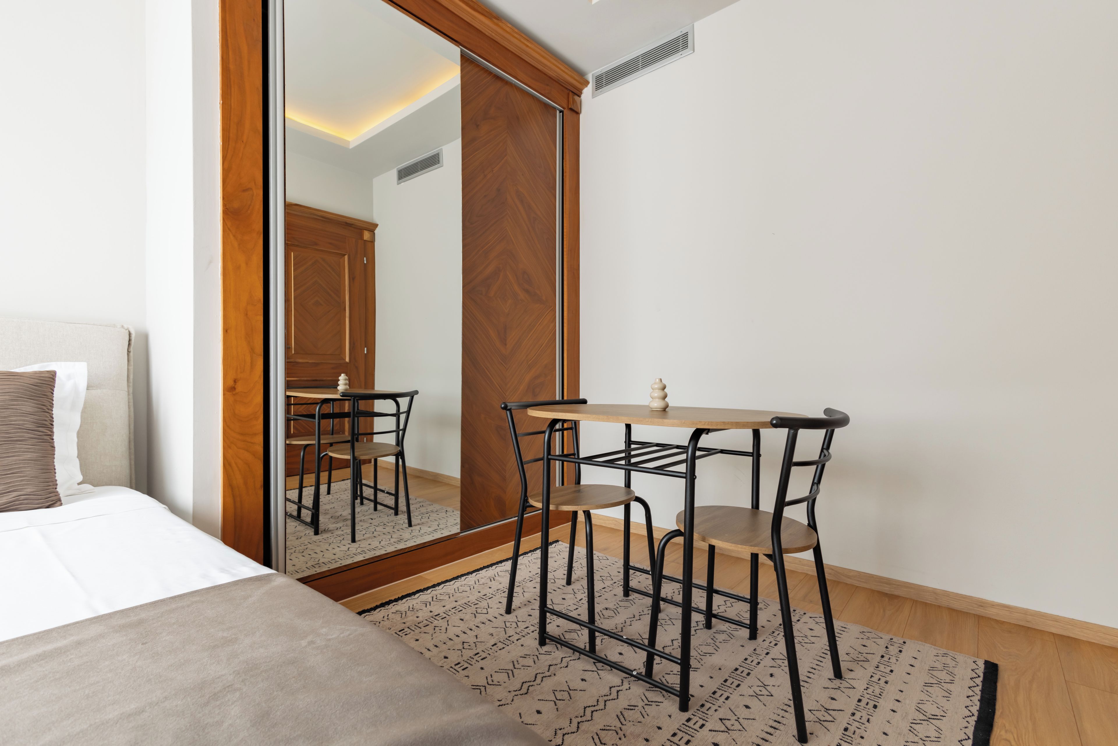 A bedroom with a bed, a desk with chairs, a mirror, and a patterned rug.