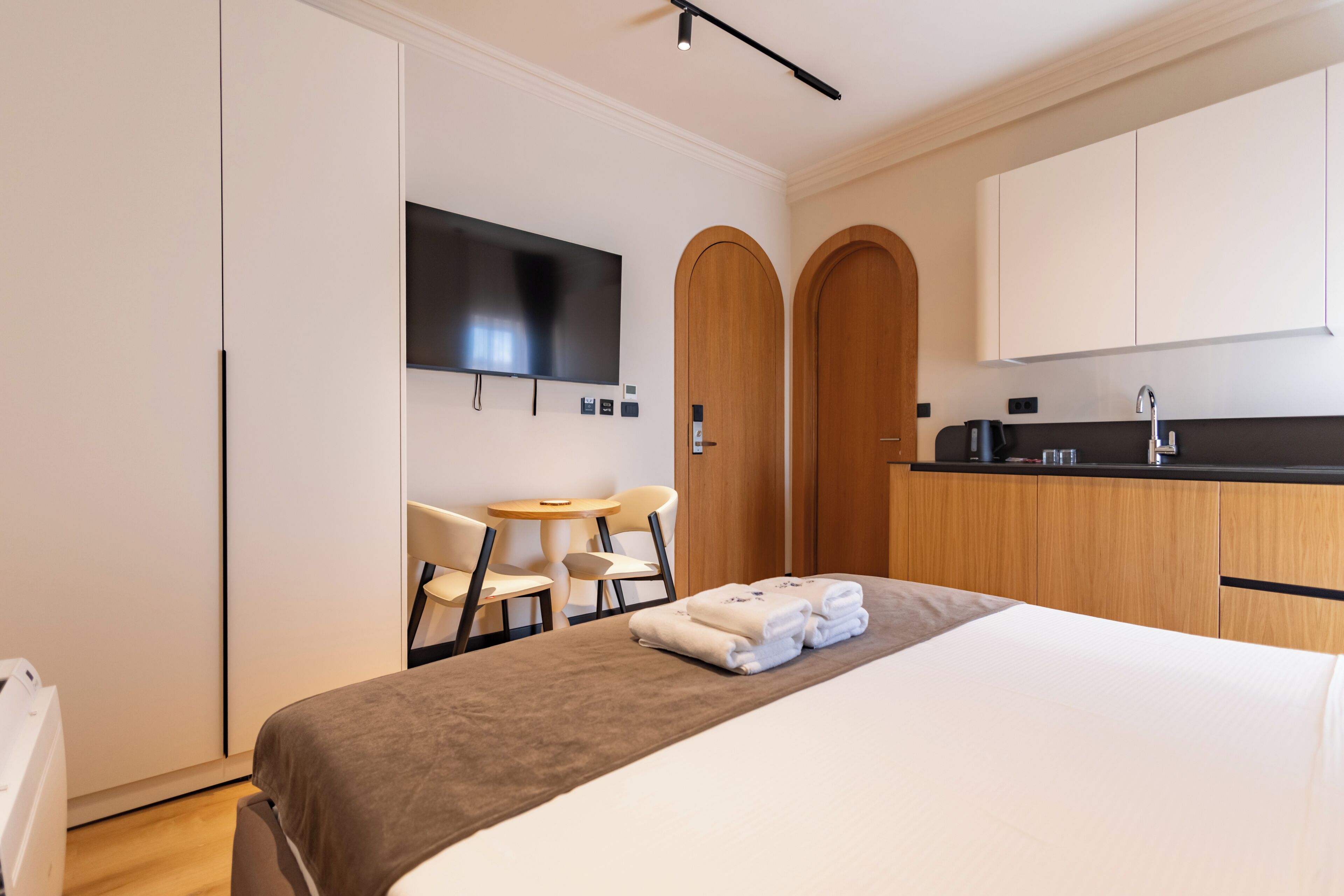 A hotel room with a bed, a desk with chairs, a flat-screen TV, a wooden wardrobe, and a kitchenette.