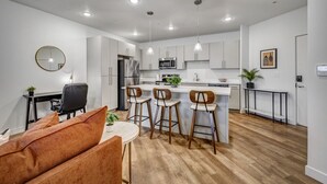 Een moderne keuken met witte kasten, een centraal eiland met barkrukken, een eethoek met een ronde tafel en een oranje bank, en een klein kantoor met bureau en stoel.