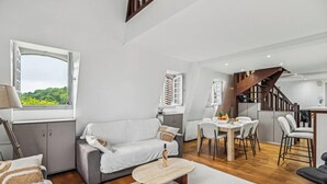 Een moderne woonkamer met een bank, eettafel en stoelen. Er is een raam met uitzicht op groen, een trap en een keukenhoek.