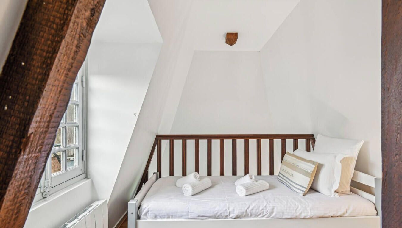 Een netjes opgemaakt bed met witte lakens en een houten hoofdbord in een kamer met witte wanden en een schuine plafond.