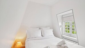 Een slaapkamer met een bed, witte lakens, een raam met jaloezieën en een bedlampje.
