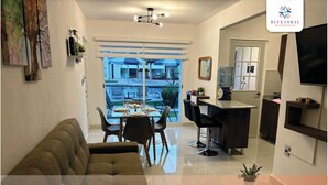 Una sala de estar moderna con un sofá, comedor con sillas y una mesa, cocina con barra y un ventanal con vistas al exterior.