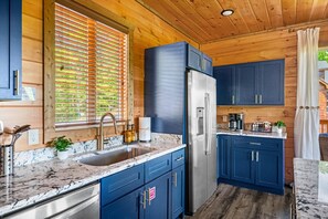 Una cocina moderna con refrigerador de acero inoxidable, armarios azules, encimera de mármol y una ventana con persianas.