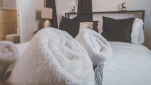Een net opgemaakt bed met witte lakens en opgerolde handdoeken, een nachtkastje met een lamp en een donker hoofdbord.