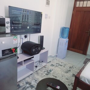 Ein Wohnzimmer mit flachbildfernseher an der Wand, einem Kühlschrank, einem Wasserspender und einer Fernbedienung auf dem Boden.