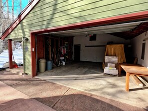 A garage with a green exterior, a red roof, and a concrete floor. There are two trash bins, a broom, and a ladder inside.