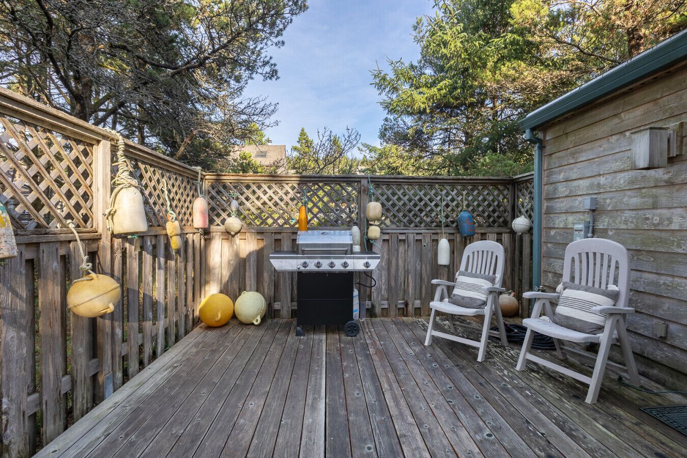 A wooden deck with a grill, two Adirondack chairs, and a wooden fence with hanging lanterns.