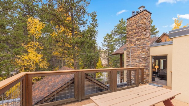 A wooden deck with a stone chimney, a wooden table, and a staircase leading to a covered area. - Columbine #301 (Breckenridge)