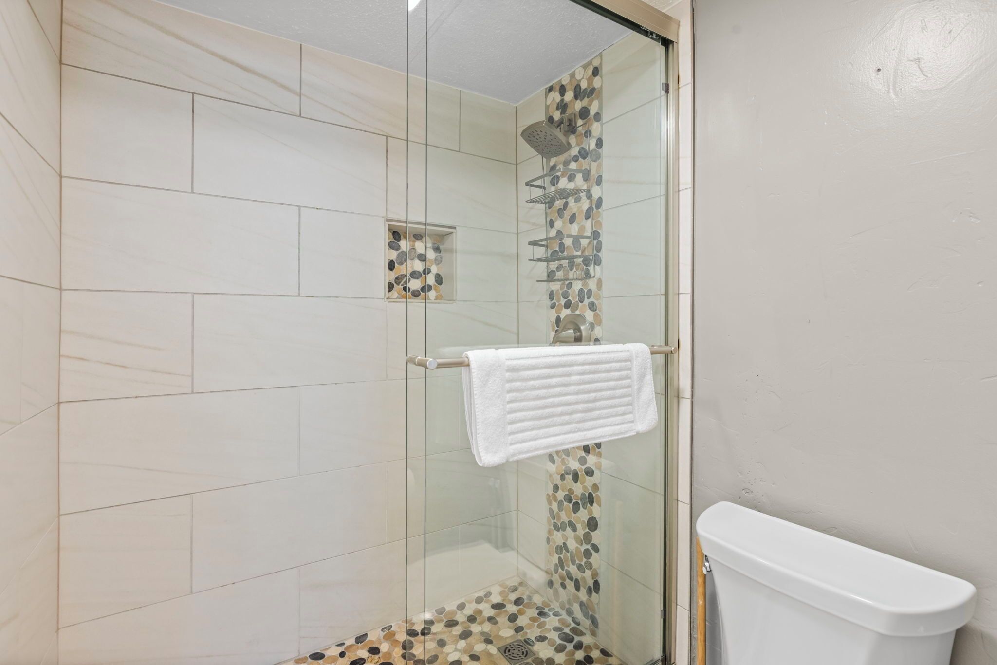 Une douche fermée par des parois en verre, avec une serviette blanche suspendue à un porte-serviettes, une porte de douche en verre et des toilettes avec une cuvette blanche.