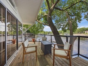 Een houten terras met een glazen balustrade, buitenmeubilair en uitzicht op een wateroppervlak.