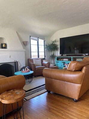 Ein Wohnzimmer mit einem braunen Ledersofa, einem blauen runden Tisch, einem Fernseher und einem großen Fenster mit Vorhängen.