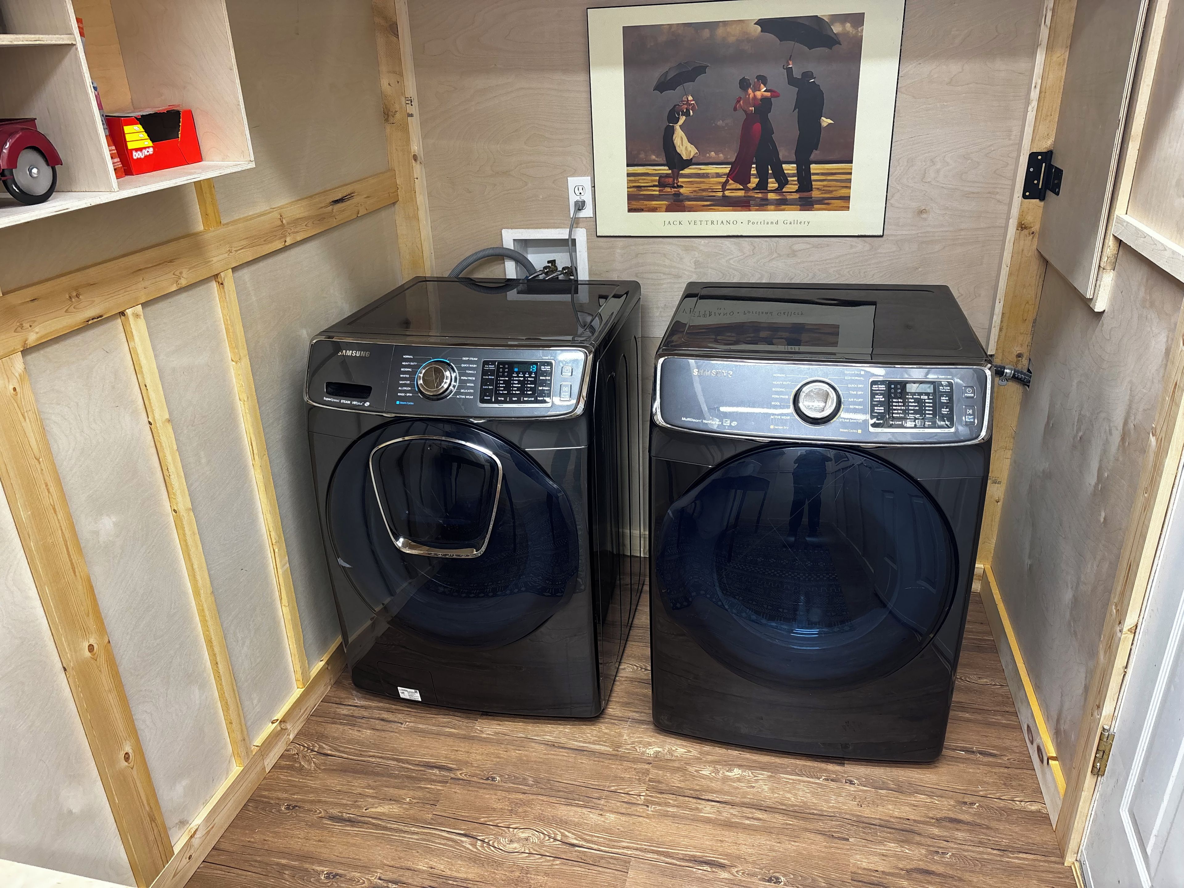 A compact laundry room with two front-loading washing machines, a power outlet, and a framed artwork on the wall.