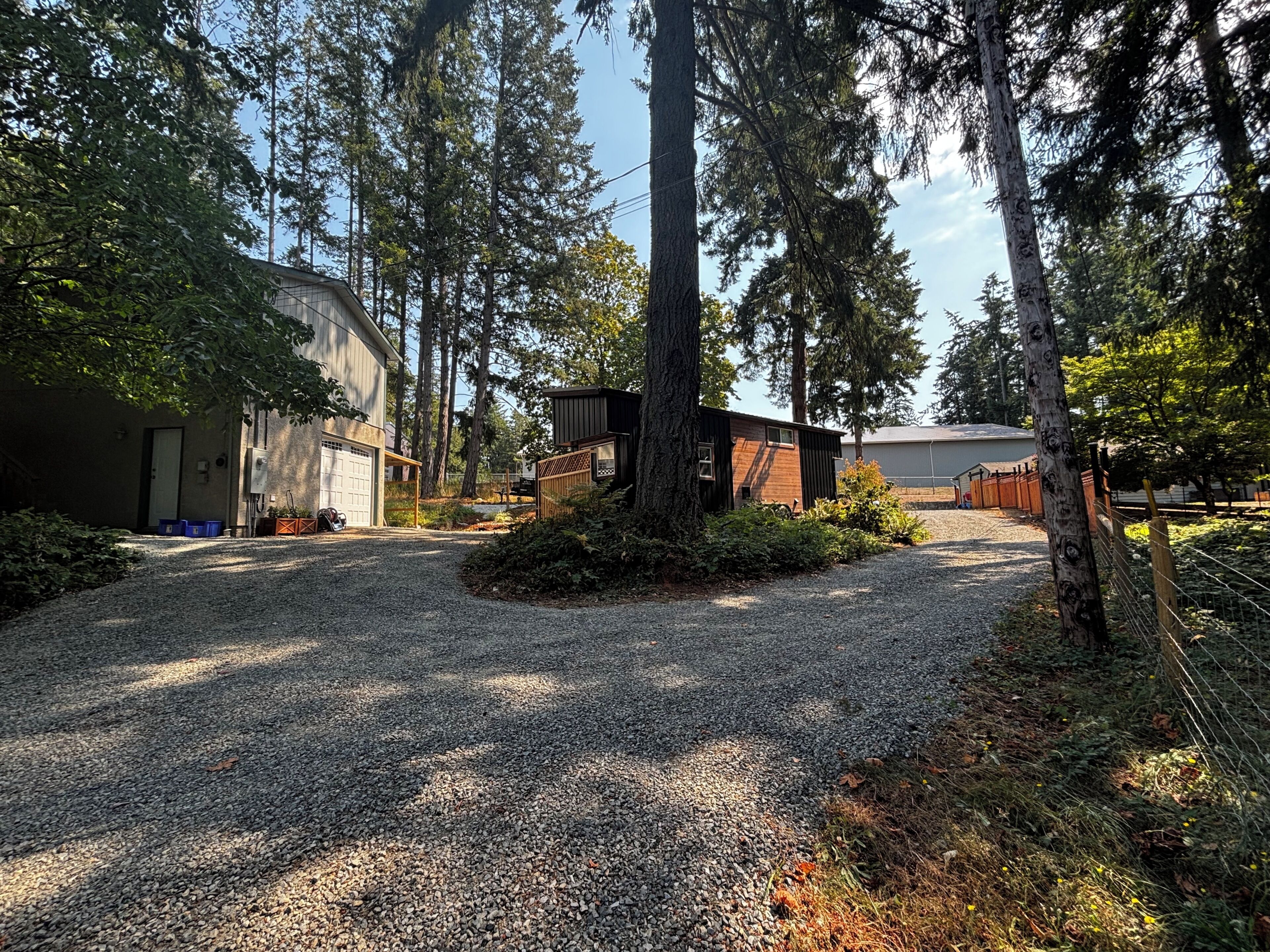 A gravel driveway leading to a building with a garage, surrounded by trees and a fenced area.