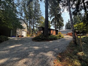Une allée de gravier mène à un bâtiment doté d’un garage, qui est entouré d’arbres et d’une zone délimitée par une clôture.