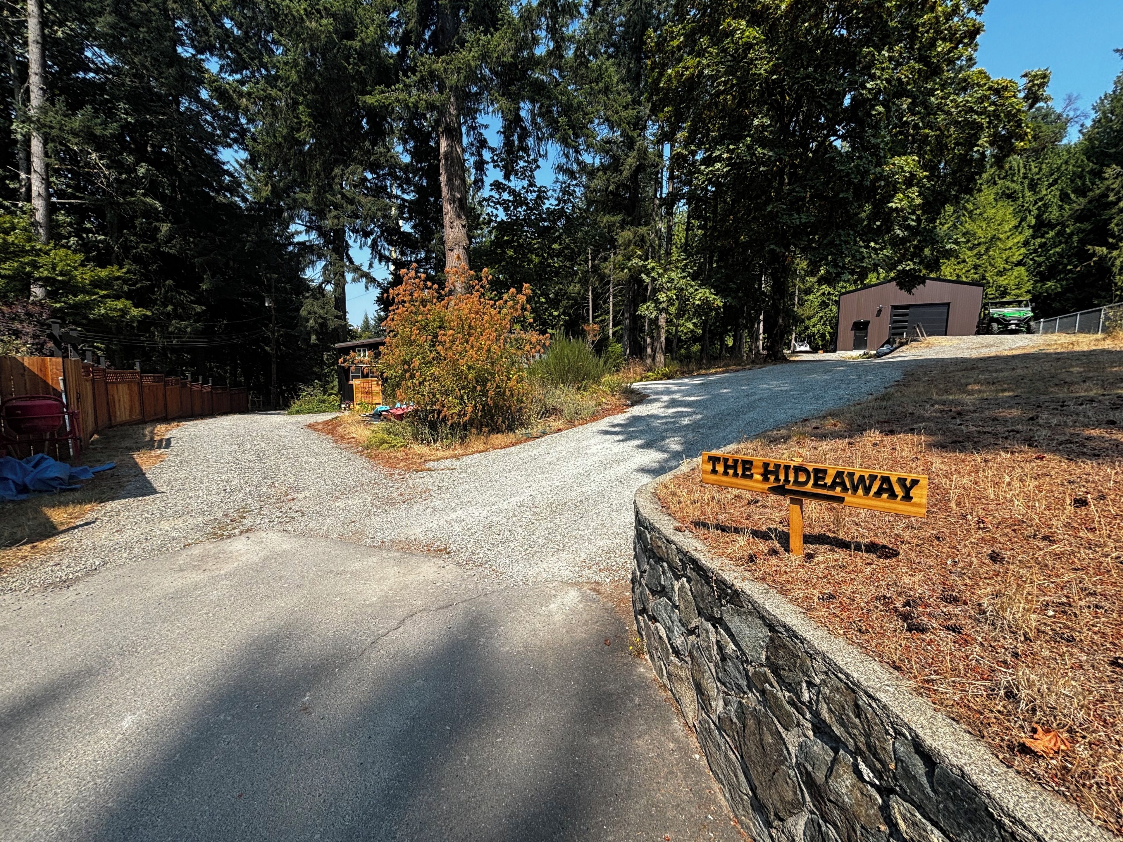 A gravel driveway leading to a wooden sign labeled 'THE HIDEAWAY' with a stone wall on one side and a building in the background.