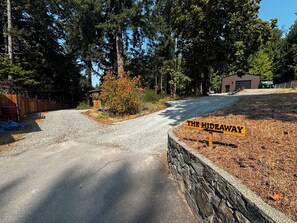 Une allée de gravier mène à un panneau en bois sur lequel on peut lire « THE HIDEAWAY ». Un mur de pierres borde l’allée d’un côté, et un bâtiment se trouve au fond.