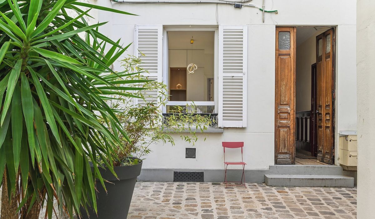 Un bâtiment doté d’une cour pavée de pierres, d’un pot de fleurs, d’une chaise rouge et d’une porte en bois.