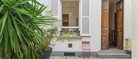 Un bâtiment doté d’une cour pavée de pierres, d’un pot de fleurs, d’une chaise rouge et d’une porte en bois.