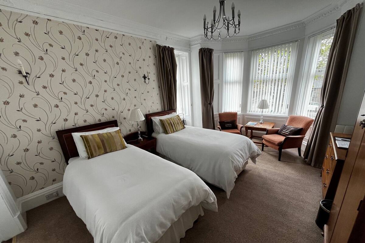 Habitación de hotel con dos camas, un candelabro, una ventana con cortinas, una silla y un escritorio.