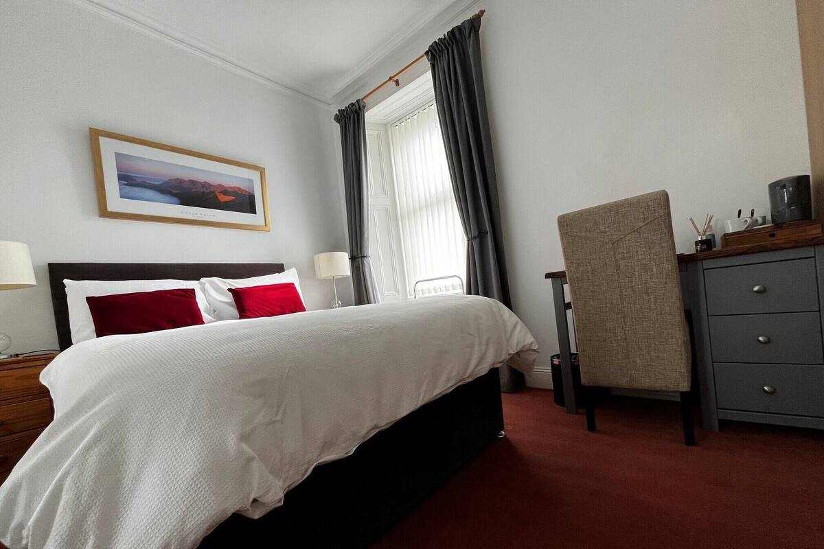 Habitación de hotel con cama, escritorio, silla, cómoda, ventana con cortinas e imagen enmarcada en la pared.