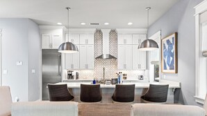 A modern kitchen with white cabinets, a stainless steel refrigerator, and pendant lighting. There is a dining area with a table and chairs. - 30A Pet Friendly House - Scissortail  (Panama City Beach)