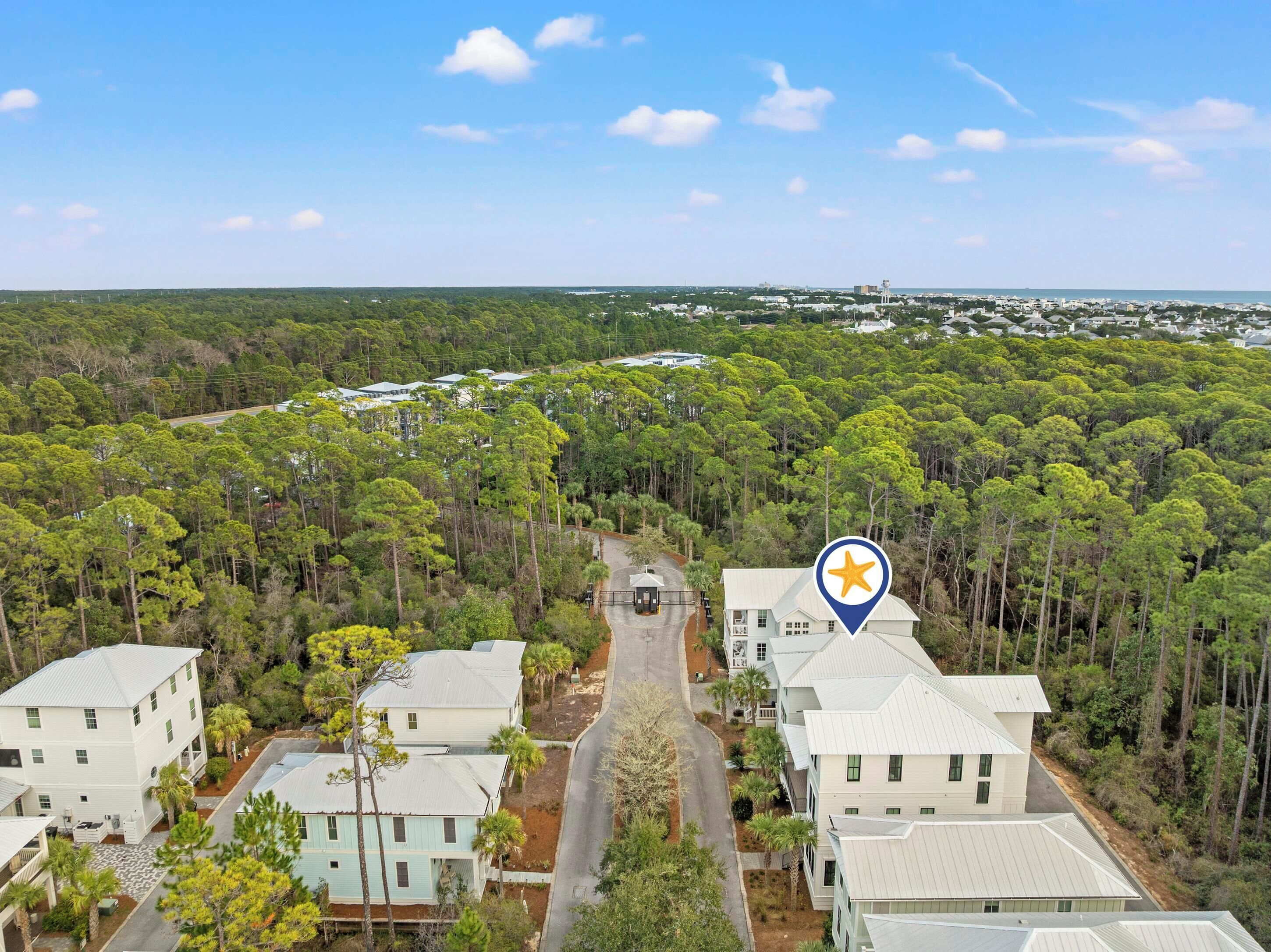 An aerial view of a residential area with houses surrounded by dense forest, a clear sky, and a distant view of a water tower.