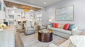 A spacious living area with a modern kitchen in the background, a round coffee table, and a large abstract painting on the wall. - 30A Pet Friendly House - Scissortail  (Panama City Beach)