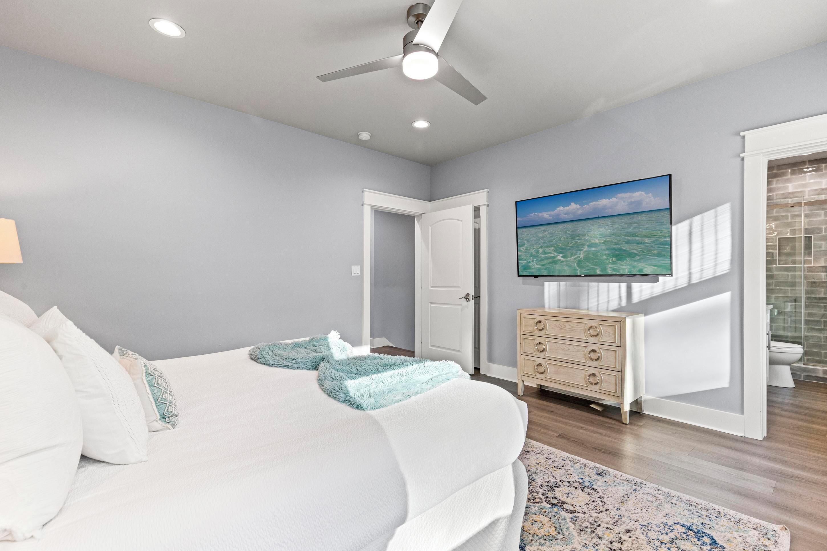 Un dormitorio con una cama, ventilador de techo, televisión, cómoda y un baño visible a través de una puerta abierta.