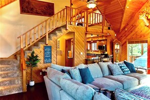 A cozy cabin interior with a wooden staircase, a living area with a sofa, and a kitchen visible through an open door. - Circle C on Timberline (Ruidoso)