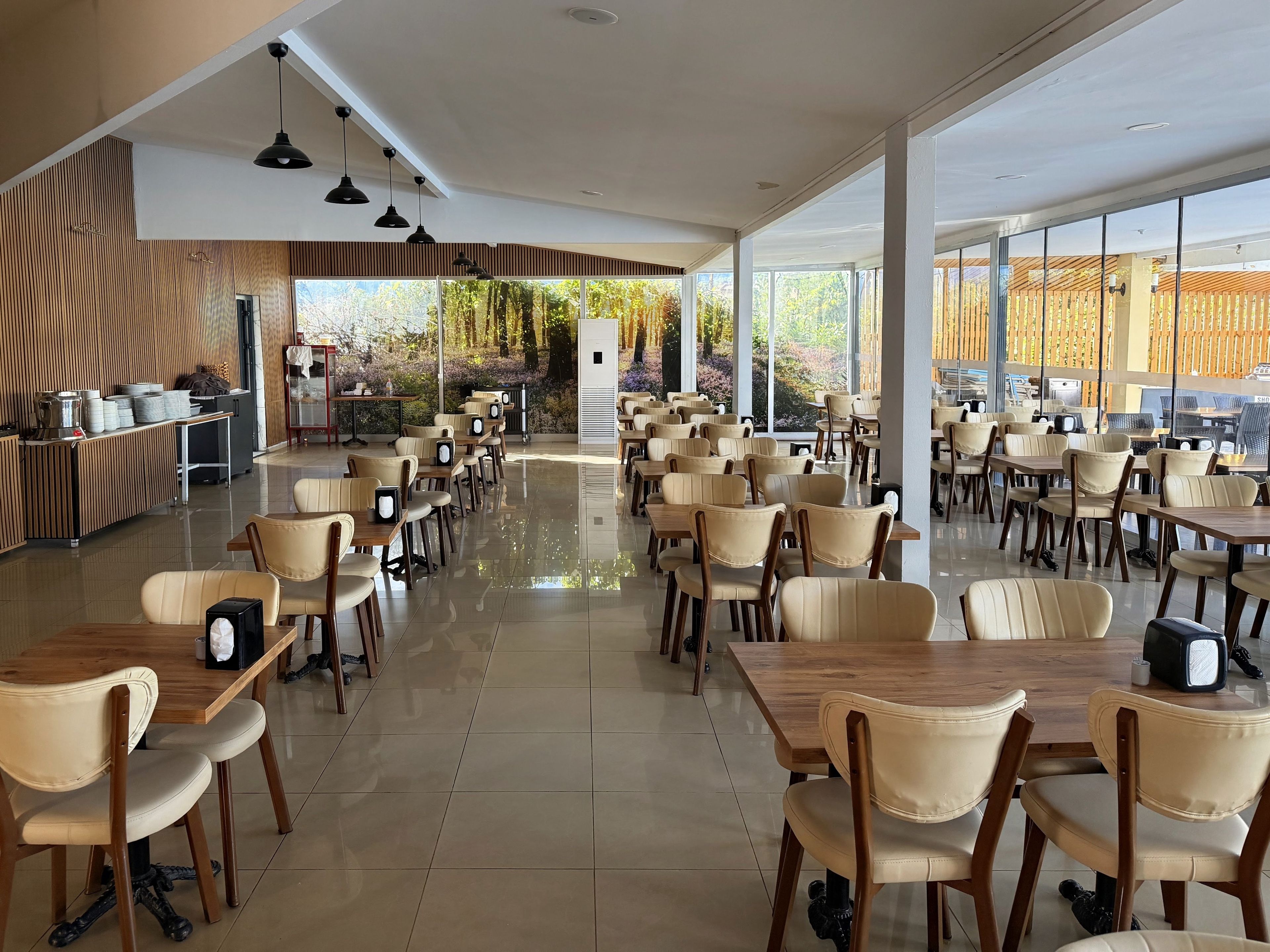 A spacious indoor dining area with multiple tables and chairs, large windows offering a view of the outdoors, and a reception desk with a counter.