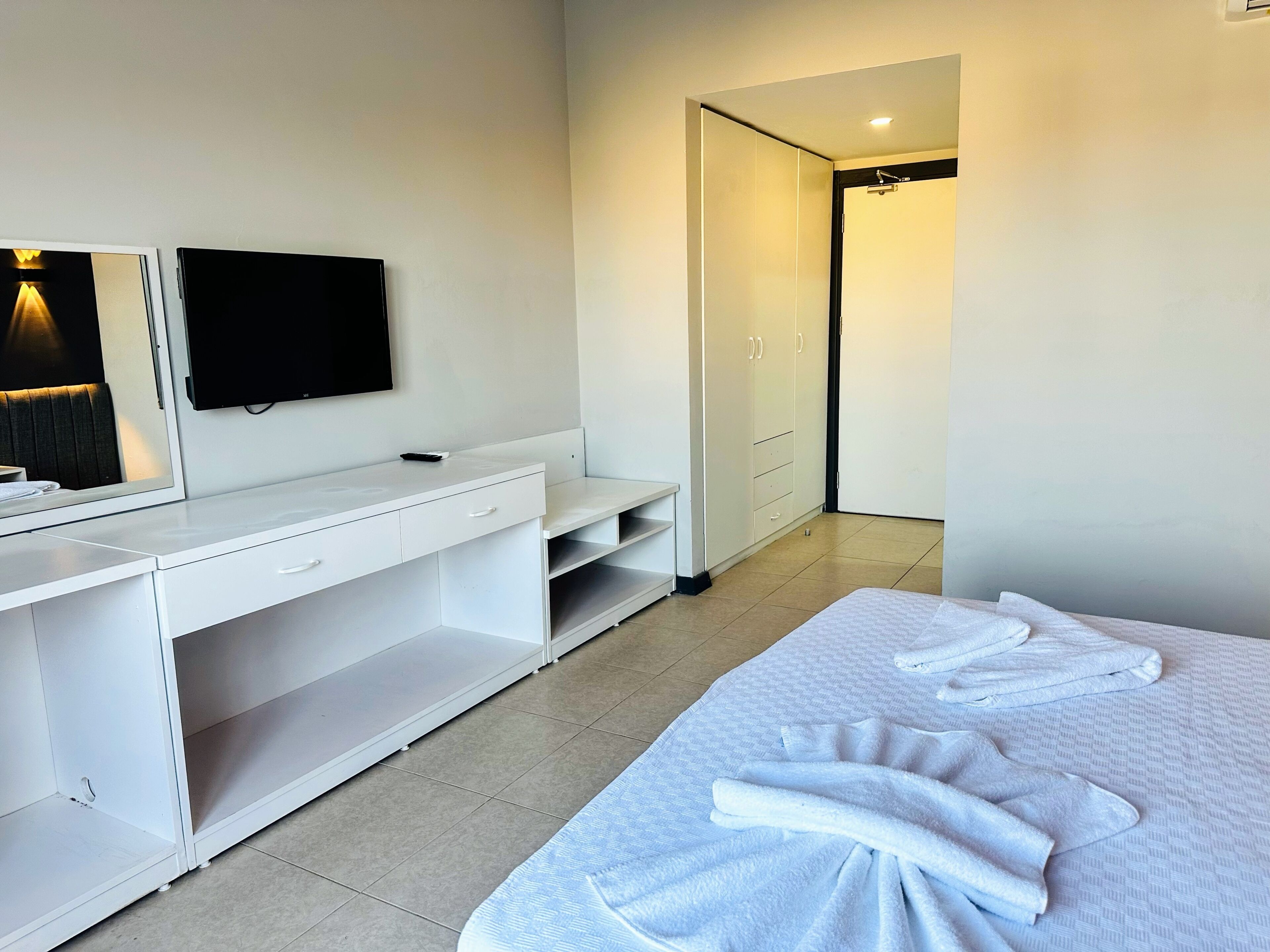 A hotel room with a bed, a desk with a flat-screen TV, a wardrobe, and a bathroom visible through an open door.
