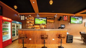 A bar area with wooden bar stools, a red refrigerator, and a television displaying a sports game. - Serenity Sports Oasis (Kampala)