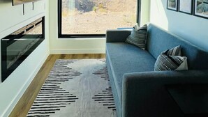 A modern living room with a large window, a flat-screen TV, a gray sofa, and a patterned rug. - Stellar Retreat at Big Bend - Cabin #8 (Terlingua)