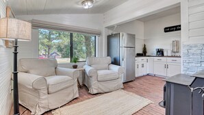Een moderne keuken met witte kasten, een roestvrijstalen koelkast en een accentmuur van steen. Er staan twee beige fauteuils en een kleine tafel met een potplantje.