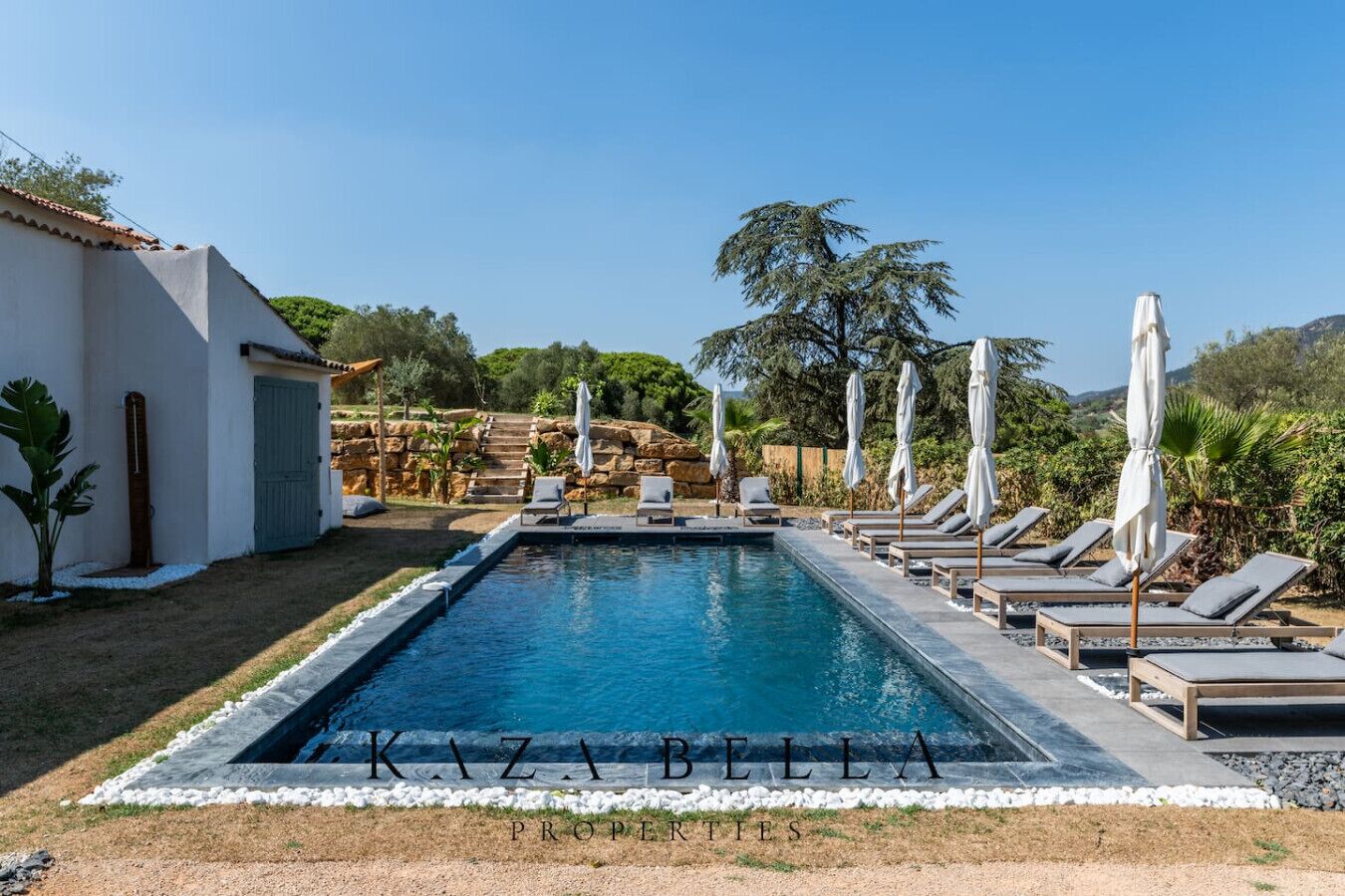 Piscina rectangular con sillas de descanso y sombrillas, rodeada por un muro de piedra y vegetación.