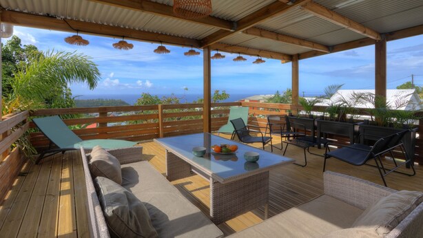 A covered outdoor seating area with wicker furniture, a glass-top table, and a view of the ocean. - Labitation - Vue sur l'océan (Saint Joseph)