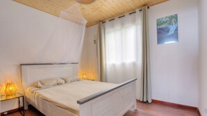 A bedroom with a wooden bed, a bedside table with a lamp, a mosquito net, and a painting on the wall. - Labitation - Vue sur l'océan (Saint Joseph)