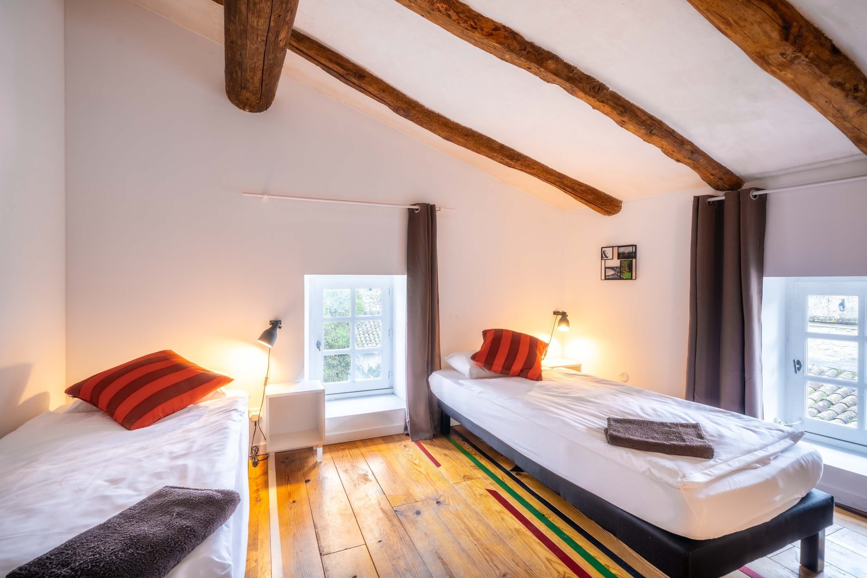 A room with two beds, wooden beams on the ceiling, a window with curtains, and a small wall-mounted picture.