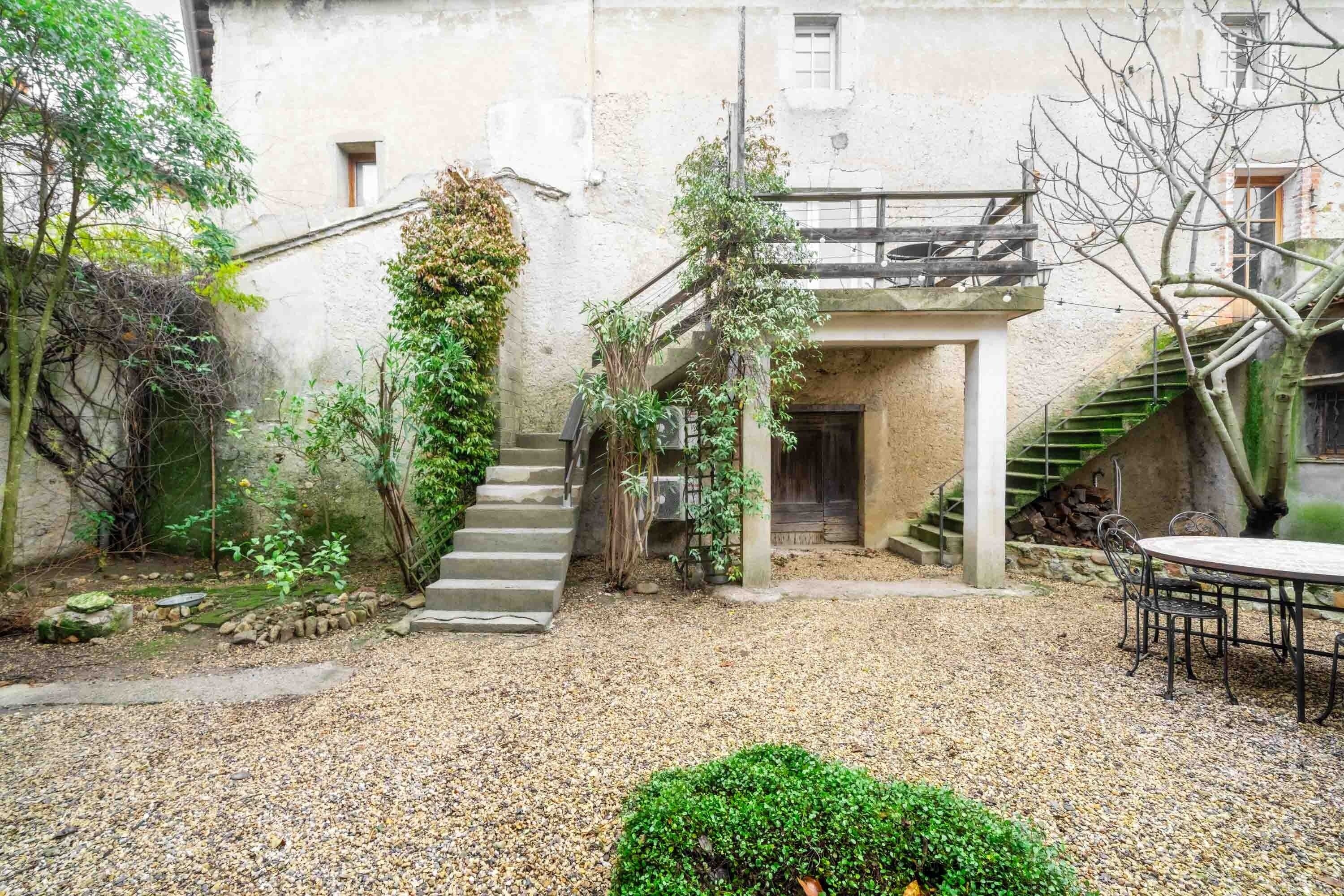 A courtyard with a gravel ground, a staircase, a small table with chairs, and a building with a balcony.