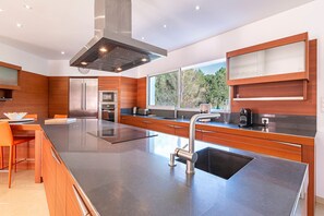A modern kitchen with a large island, stainless steel appliances, and a window with a view of trees. - Villa Maria Magdalena - Avec piscine (Sari-Solenzara)