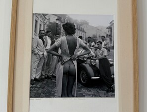 Een oude foto van een vrouw in een jurk met een diep uitgesneden rug, staand op een geplaveid straatje met een klassieke auto en toeschouwers.