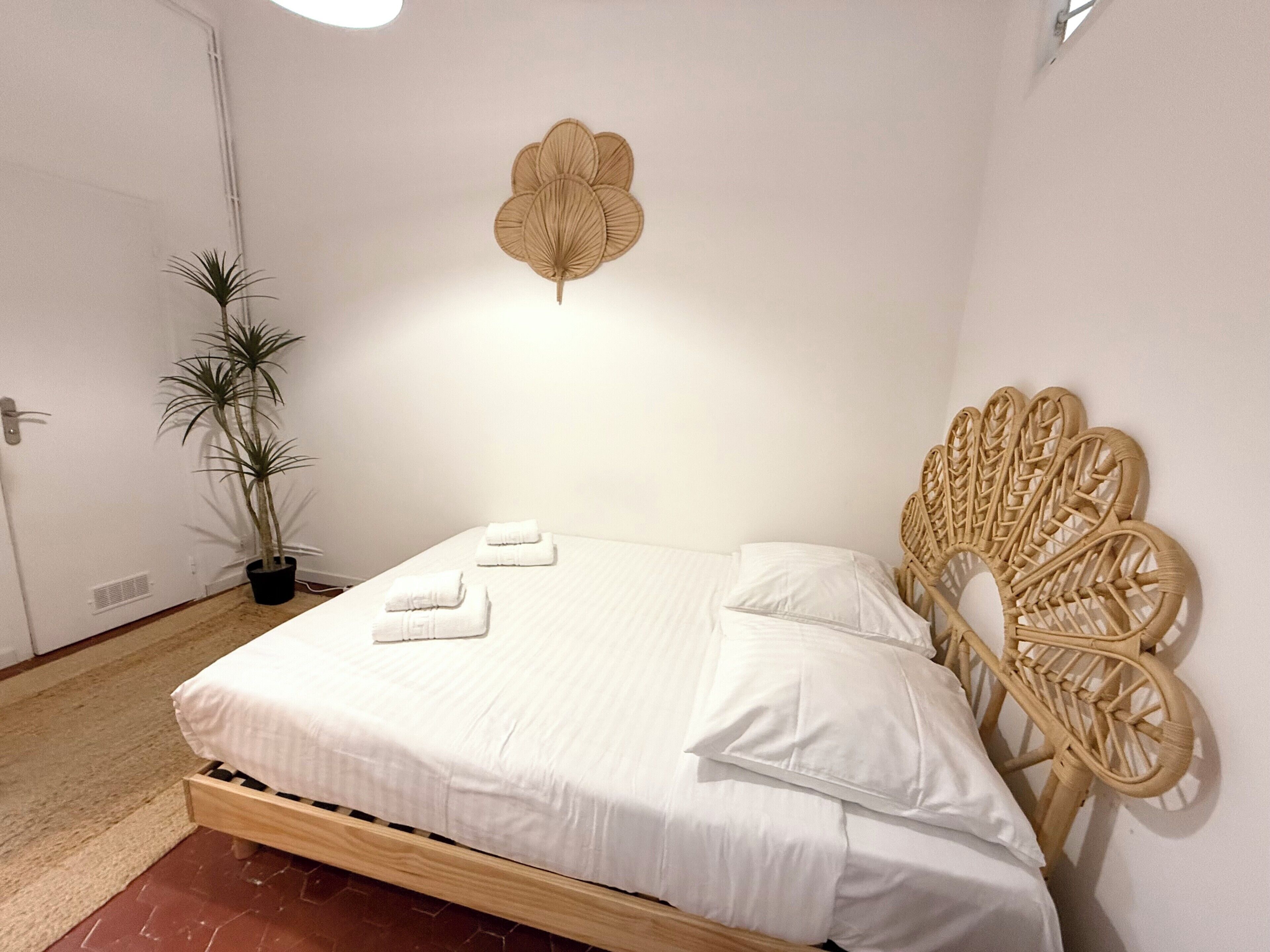 Una cama bien hecha con sábanas blancas, una planta en maceta a su lado y una hoja decorativa en la pared.