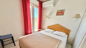 Ett hotellrum med en säng, ett litet bord, en stol, ett fönster med röda gardiner, en luftkonditioneringsanläggning och en vägglampa.