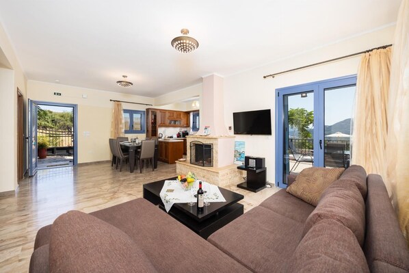 A living room with a sofa, a coffee table with a bottle, and a TV. There is a kitchen in the background with wooden cabinets and a dining area.