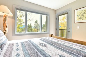 Ein Schlafzimmer mit großem Fenster, einem Bett mit gemustertem Überwurf, einem Nachttisch und einer Tür mit Blick auf Bäume.