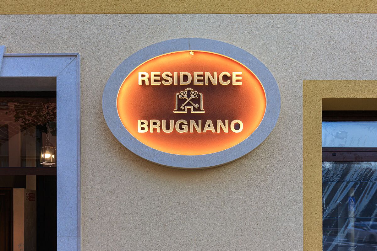 Un letrero circular con la leyenda 'RESIDENCE BRUGNANO' y un logotipo, colocado en una pared de edificio.
