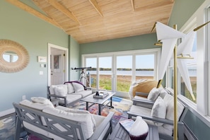 A room with a wooden ceiling, a large window with a view of the beach, a white sofa, a small table, and a chair. - Wonderland on the Water with Exclusive Beach Access (Biddeford)