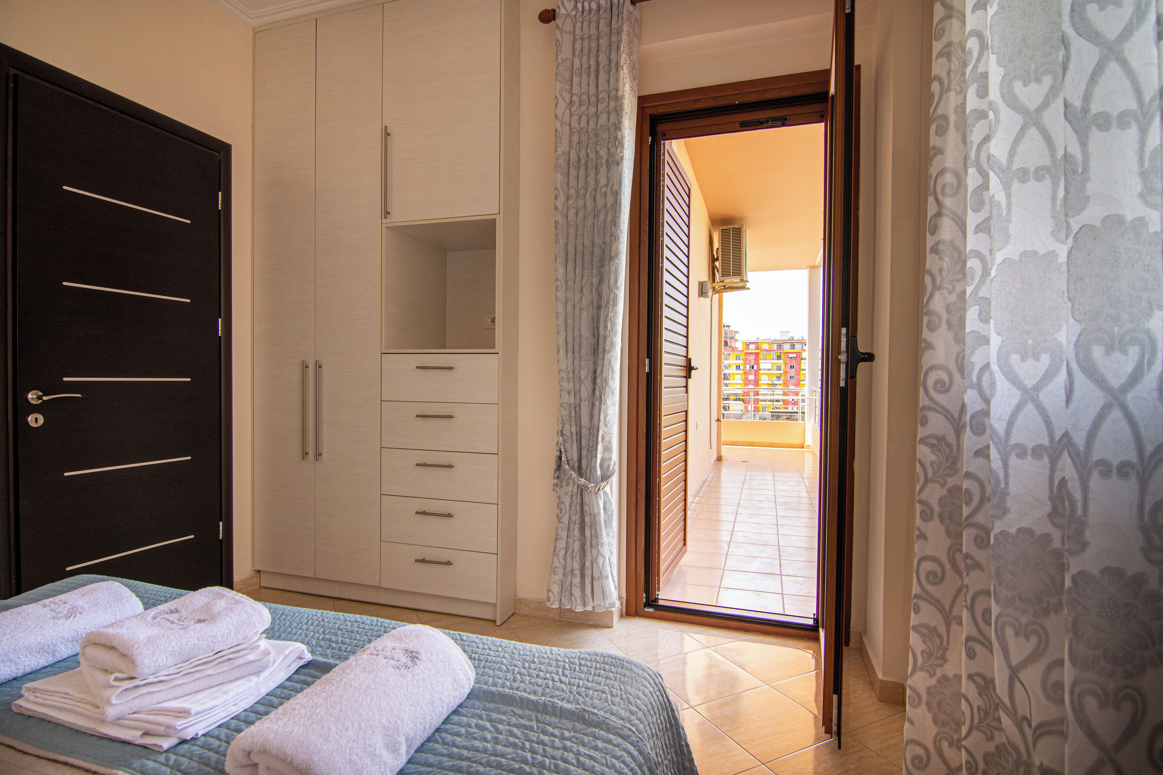 A bedroom with a bed, a wardrobe, a bathroom visible through an open door, and a view of the outside.